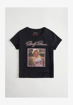 Camiseta negra de manga corta con una mujer sonriente con vestido vintage y una flor en el pelo, y texto estilizado sobre la imagen.