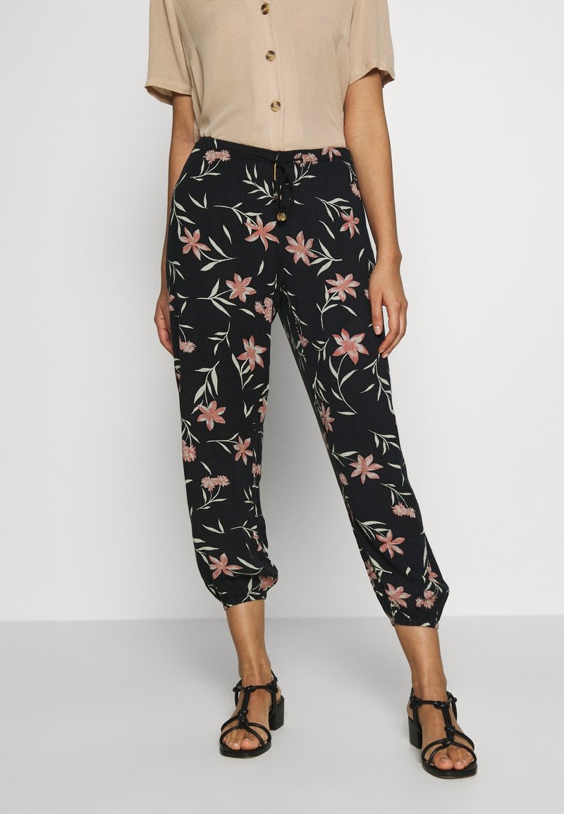 Femme portant un pantalon noir à imprimé floral avec cordon de serrage, un haut beige à manches courtes boutonné et des sandales à talons noires avec brides, debout contre un mur uni.