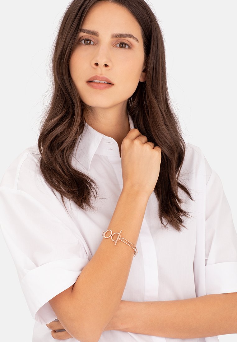 Rose gold bracelet with a toggle clasp, featuring an elongated link design. The model wears a white shirt with rolled sleeves, showing the accessory.