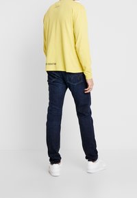 Camisa de algodón amarilla de manga larga con texto en la parte de atrás, combinada con jeans de mezclilla azul oscuro y zapatillas blancas, vista desde la parte trasera.