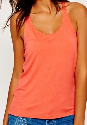 Woman wearing coral sleeveless tank top with lace detail on straps and blue denim jeans against a white background.