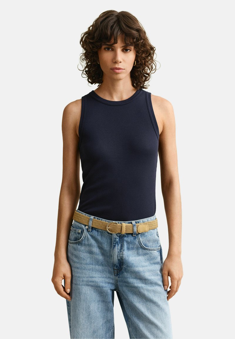 Femme aux cheveux bruns bouclés, portant un haut sans manches bleu marine, un jean bleu clair et une ceinture tressée beige, debout devant un fond uni.