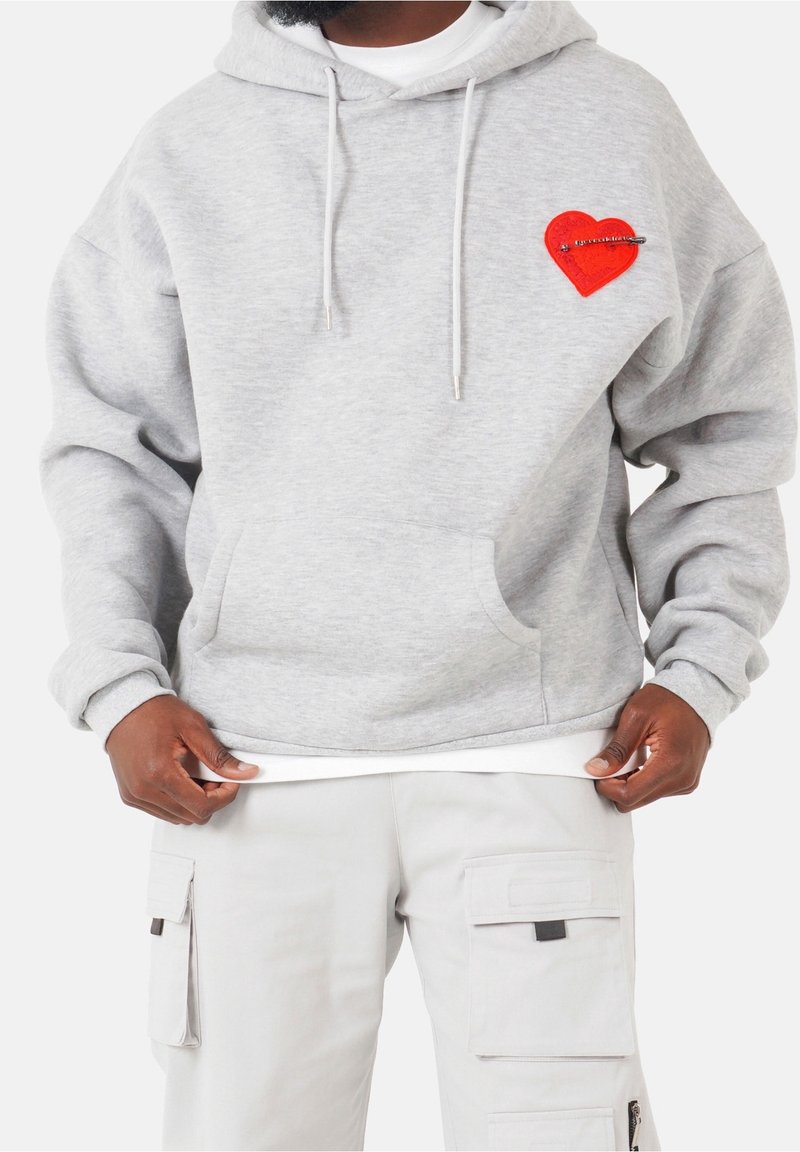Homme portant un sweat à capuche gris clair avec un patch cœur rouge épinglé sur la poitrine, associé à un pantalon cargo clair à multiples poches.