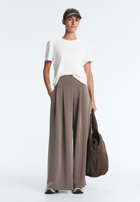 Femeie îmbrăcată într-un tricou alb, pantaloni largi taupe, șapcă neagră, adidași, ținând o geantă mare de culoare maro, pe un fundal simplu.