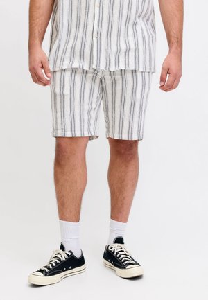 Homme portant un short rayé blanc et gris et une chemise à manches courtes assortie, baskets noires et chaussettes blanches, debout sur fond blanc.