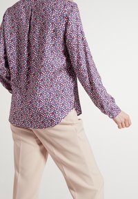 Blouse fleurie violette, avec un motif de petites fleurs roses, bleues et blanches, manches longues et ourlet arrondi. Associée à un pantalon beige.