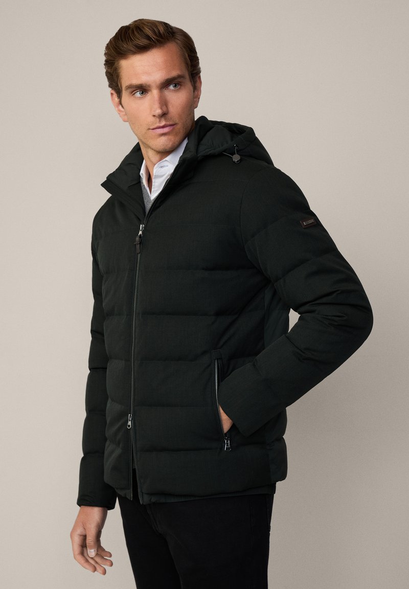 Uomo che indossa un giubbotto di piuma nero con cappuccio, dotato di tasche con zip, e una camicia chiara sotto, che guarda a sinistra su uno sfondo semplice.