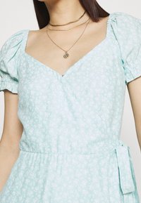 Himmsk blå blomsterinspirerad wrapklänning med puffärmar och v-ringning. Har en åtsittande midja och detalj med sidoknytnig, tillverkad av lättviktsmaterial.