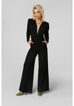 Donna con capelli biondi che indossa una tuta nera con scollo a V profondo, maniche lunghe e gambe larghe, abbinata a décolleté verdi oliva con punta affilata.