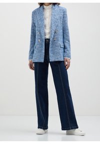 Blazer texturé bleu avec fermeture croisée à double bouton doré, associé à un pantalon en denim foncé taille haute et des baskets blanches.