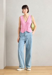 Rosa stickad väst med v-ringning, framknappar och fickor. Tillsammans med ljusblå vida jeans och vita sneakers, kompletterat med en grön väska.