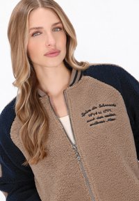 Fleece bomberjakke med en tan farve på kroppen, marineblå ærmer, ribbet krave og broderet tekst på forsiden. Detaljer om lynlåslukning vises.