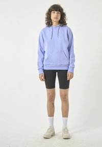 Hellblaues Kapuzensweatshirt mit einer Vordertasche, getragen über schwarzen Radsportshorts. Das Modell trägt weiße Socken und beige Turnschuhe.