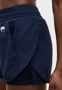 Κοντινή λήψη ατόμου που φοράει αθλητικό σορτς σε ναυτικό μπλε χρώμα με επικαλυπτόμενα πλαϊνά πάνελ και λευκό λογότυπο φιδιού στην αριστερή πλευρά.