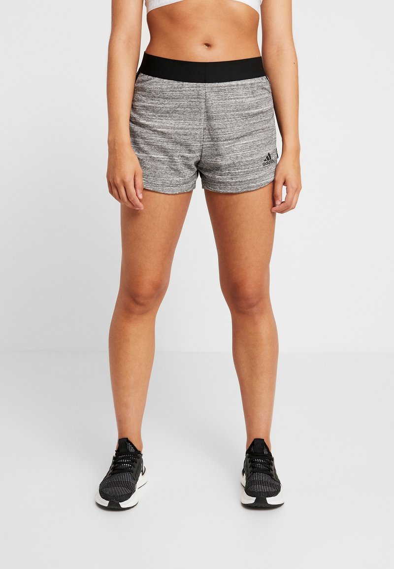 Femme portant un short de sport Adidas gris avec une ceinture noire et des chaussures de course noires, debout sur un fond blanc uni.