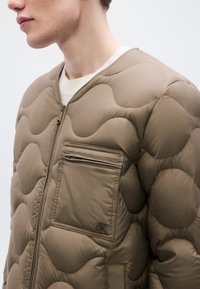 Ανοιχτό καφέ καπιτονέ μπουφάν με στρογγυλή λαιμόκοψη, κλείσιμο με φερμουάρ μπροστά και τσέπη στο στήθος με φερμουάρ. Διαθέτει υφασμάτινη, επενδυμένη επιφάνεια με υφή και λεπτομέρεια με λογότυπο.