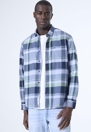 Camisa de franela a cuadros en azul, verde y gris. Con botones, mangas largas y un bolsillo. Llevada sobre una camiseta blanca.