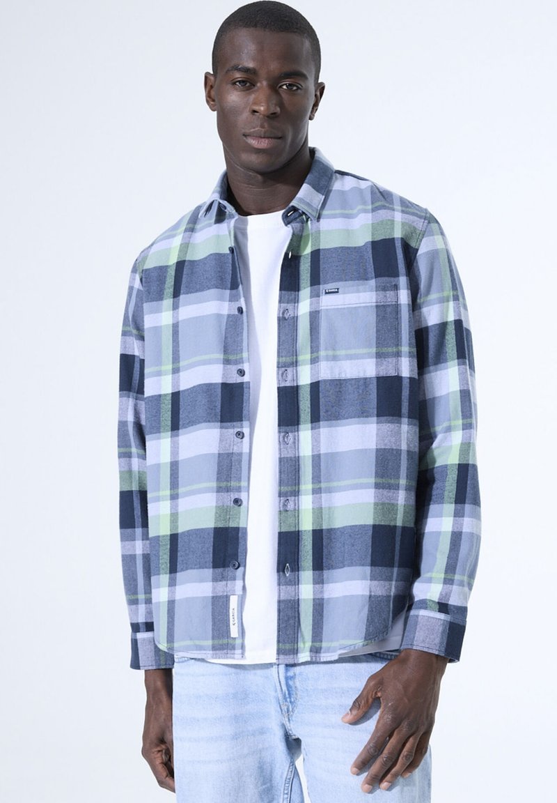 Geruite flanellen shirt in blauw, groen en grijs. Heeft knopen, lange mouwen en een zak. Draagt over een wit T-shirt.