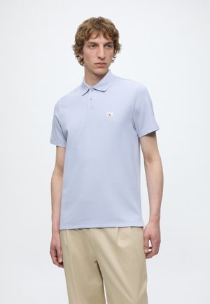 Ung mand med krøllet hår iført en lyseblå polo med logo og beige bukser mod en ensfarvet hvid baggrund.