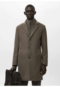 Abrigo de lana marrón con cuello estructurado, cierre de cinco botones, bolsillos laterales y textura suave. La modelo sostiene un bolso oscuro de ante.