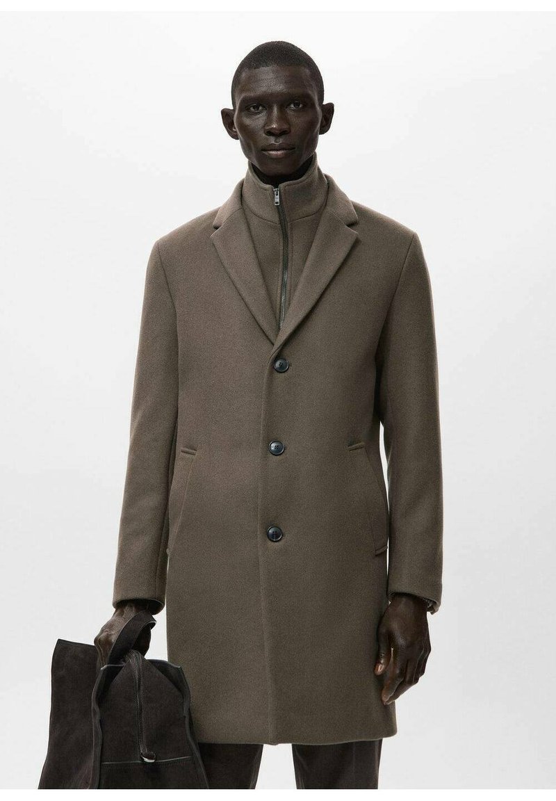 Abrigo de lana marrón con cuello estructurado, cierre de cinco botones, bolsillos laterales y textura suave. La modelo sostiene un bolso oscuro de ante.