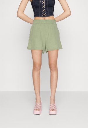 Femme portant un short vert clair à taille élastique, un crop top bleu foncé à lacets et des sandales compensées roses avec des brides à carreaux.