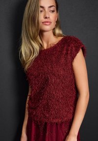 Burgundy yläosa, jossa on teksturoitu, fringe-muotoilu, leveät hihat ja rento istuvuus, yhdistettynä sopivaan brodeerattuun hameeseen.