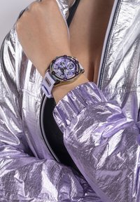 Lilla analogt ur med en krystaldekoreret bezel, hvid silikonerem, og en struktureret sølvfarvet lynlåsjakke med reflekterende finish.