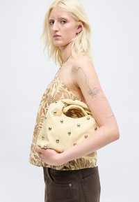 Femme blonde portant un haut à motifs et un pantalon foncé tient un sac à main jaune pâle orné de clous argentés sur fond blanc.