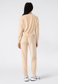 Beżowy dres z wysokim kołnierzem; gładki materiał, dopasowany krój. Połączony z białymi i czarnymi sneakersami z różowymi akcentami.