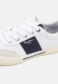Zapatilla de lona blanca con panel de tela azul marino, suela de goma, puntera de ante, y ojales de metal. Cuenta con la marca "Pepe Jeans London".