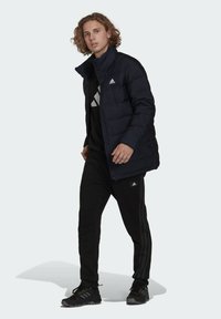 Giacca imbottita blu navy con colletto alto, chiusura a zip e logo Adidas. Indossata sopra un felpa nera e pantaloni della tuta neri con tre strisce.