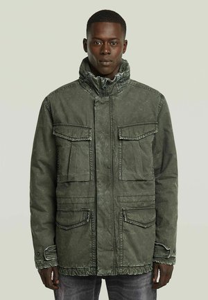 Homme portant une veste vert olive avec quatre grandes poches avant et un col montant, debout devant un fond clair uni.