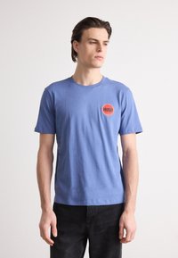 Blue cotton T-shirt featuring a round neckline and short sleeves, with a red circular logo labeled "HUGO" on the left chest.