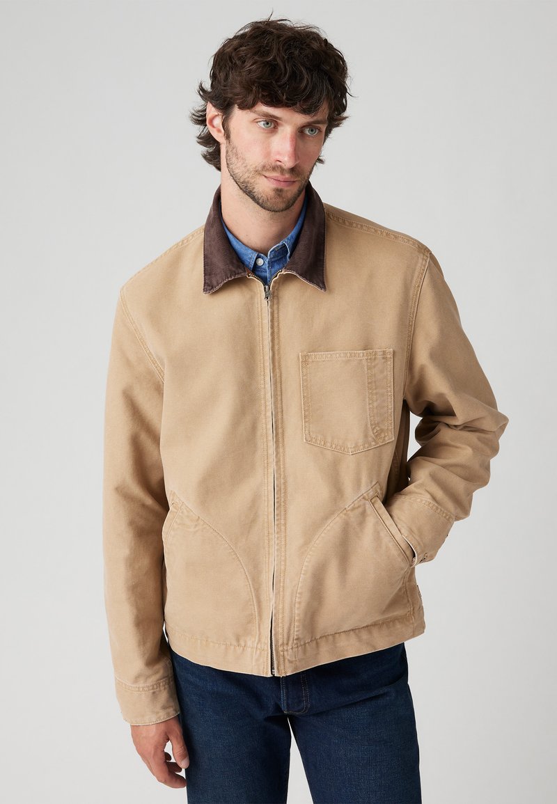 Chaqueta de algodón beige con un cuello de pana marrón, bolsillos delanteros y cierre de cremallera. Presenta una textura suave y un ajuste entallado.