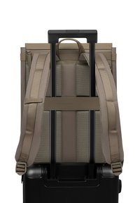 Beige stoffen rugzak met verstelbare banden, geplaatst op een zwarte koffer met wielen. Metalen handvat steekt uit de koffer.