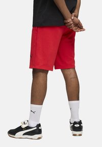 Pantalones cortos deportivos rojos hechos de una tela suave, con un logo negro en el costado. El modelo lleva zapatillas negras y calcetines deportivos blancos.