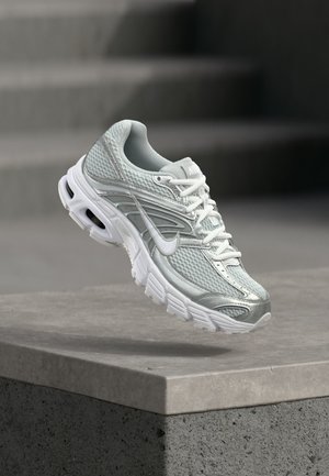 Svijetlosiva i bijela Nike tenisica za trčanje s mrežastim materijalom i debelim potplatom lebdi iznad betonskog ruba na zamućenom pozadinskom prikazu stepenica.