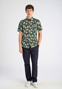 Camicia verde a maniche corte con fiori arancioni e bianchi, abbinata a pantaloni blu navy e sneakers bianche. Vestibilità casual e design con bottoni.
