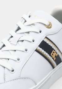 Sneaker sintetica bianca con superficie texturizzata, occhielli blu e accenti a strisce nere e oro. Presenta lacci bianchi e dettagli del logo dorato.