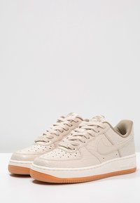 Ett par låga beiga Nike Air Force 1-sneakers med texturerad ovandel, perforerat tåparti och sula i gummi, visade sida vid sida.