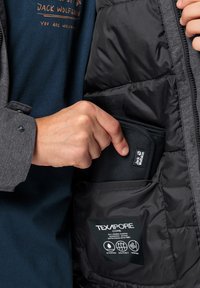 Giacca Texapore nera con una tasca interna che contiene un piccolo sacchetto nero. Mano che si infiltra nella tasca. Camicia grigia sotto la giacca.