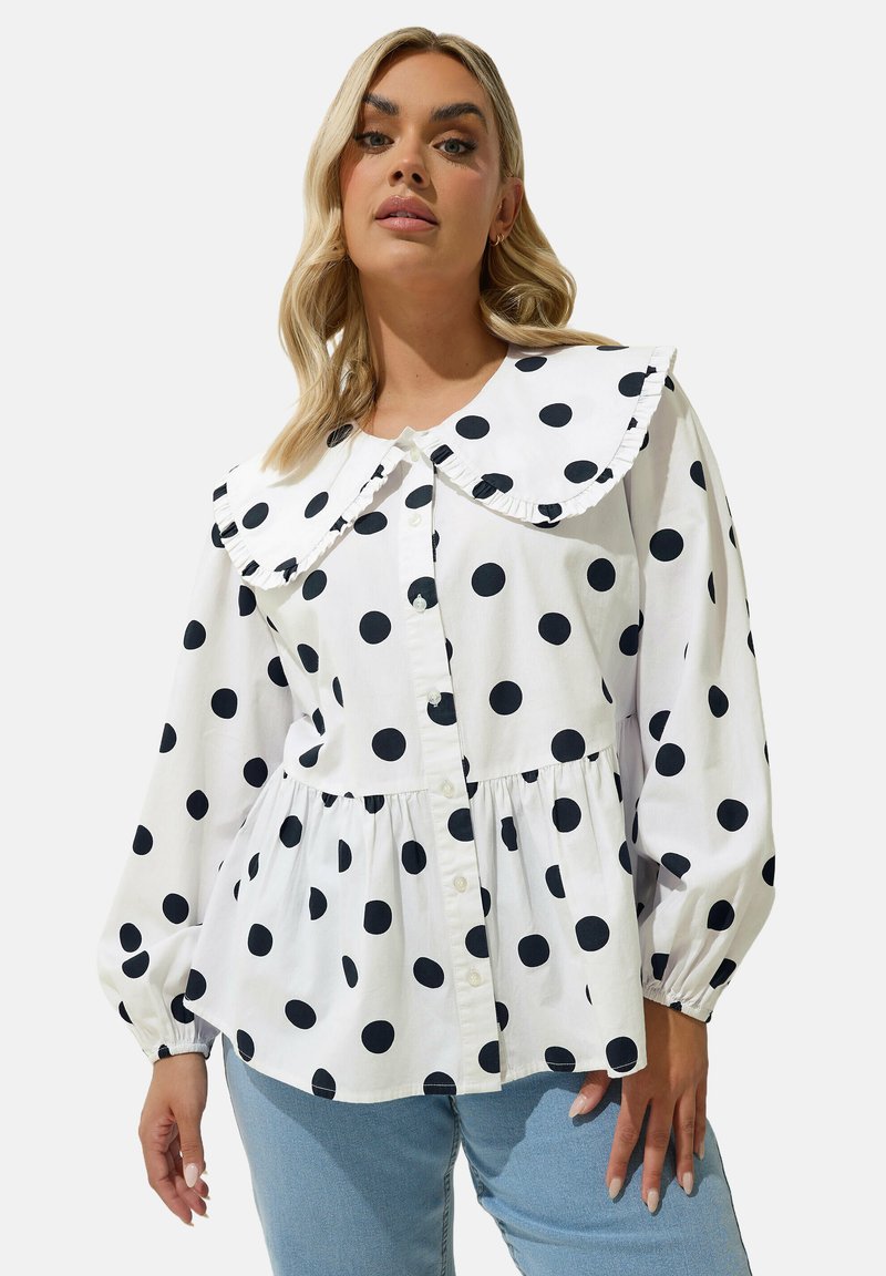 Camicia bianca con bottoni, decorata con grandi pois neri, colletto ampio e arricciato, e vita arricciata, abbinata a jeans azzurri chiari.