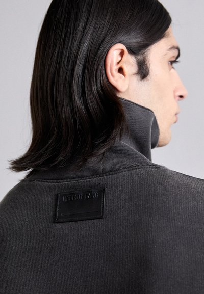 Grå høyhalset sweatshirt med ribbestruktur og et svart logomerke bak, som fremhever et rent, minimalistisk design.