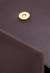 Fioletowa skóra z wyraźnym okrągłym złotym zapięciem; gładka faktura i staranne przeszycie widoczne na krawędzi.