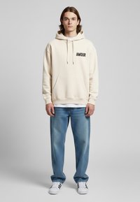 Felpa crema con testo "AMOUR" nero, tasca a marsupio, cappuccio con cordino; abbinata a jeans azzurri e sneakers bianche con strisce nere.