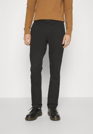 Zwarte op maat gemaakte broek met een slim fit, gemaakt van een gladde stof, met zijzakken en een knoopsluiting. Gecombineerd met glanzende zwarte schoenen.