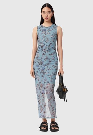 Vrouw draagt mouwloze, lichtblauwe bloemen maxi-jurk, houdt een zwarte handtas vast en draagt zwarte sandalen, staat tegen een effen achtergrond.