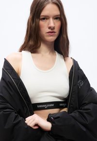 Νεαρή γυναίκα με μακριά καστανά μαλλιά φοράει λευκή κοντή μπλούζα με την επιγραφή "tommy jeans" και ένα μαύρο μπουφάν με μερικώς κλειστό φερμουάρ, ποζάροντας μπροστά σε λευκό φόντο.