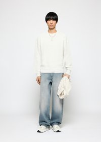 Junger Mann mit schwarzem Haar, der einen weißen Strickpullover, hellblaue weite Jeans und weiße Sneaker trägt und in seiner rechten Hand eine weiße Jacke hält.